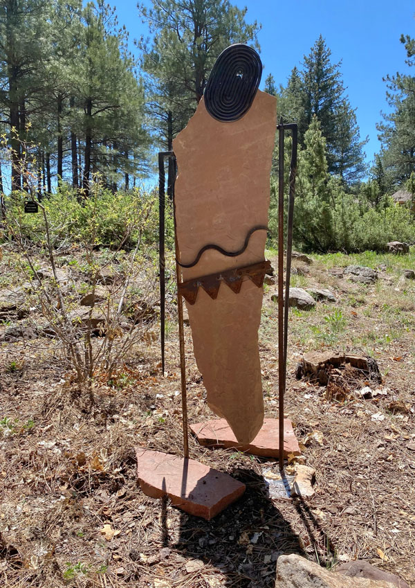 A sandstone and forged steel sculpture of a totemic figure installed in a garden at the Flagstaff Arboretum